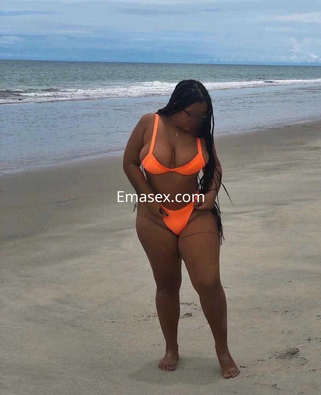 Escort en Huelva con piel oscura y trenzas, vestida con un bikini naranja, posando en la playa.