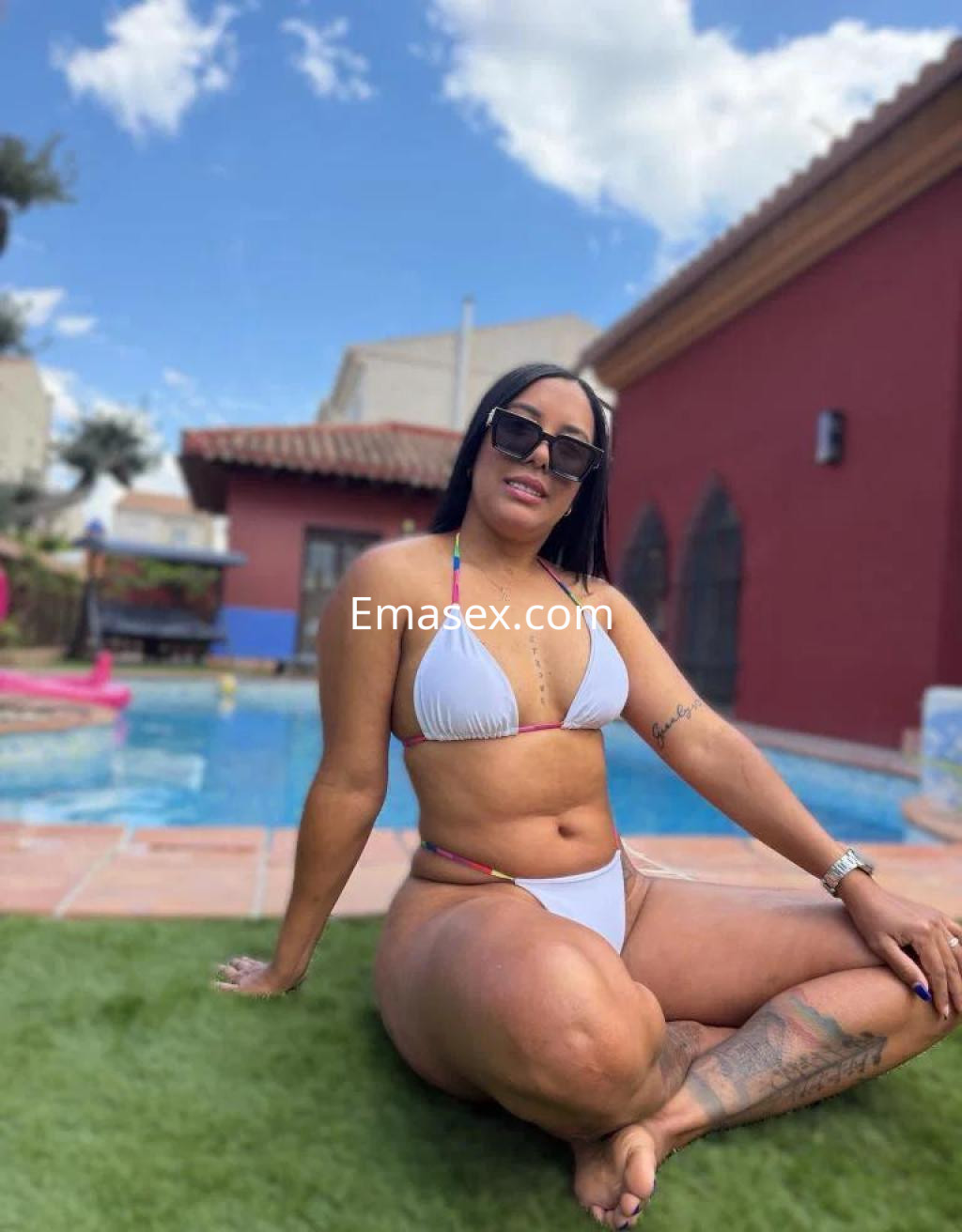 Escort en Huelva, sentada sobre el césped junto a una piscina, luciendo un bikini blanco y gafas de sol.
