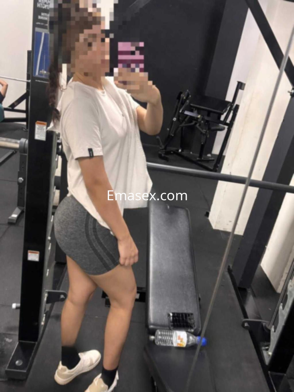 La puta en Huelva se muestra en el gimnasio, vistiendo una camiseta clara y shorts ajustados, posando frente al espejo.