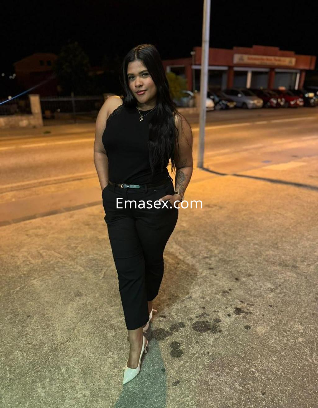 Escort en Lugo con cabello largo y oscuro, vestida con blusa negra y pantalones ajustados, posando en la calle.