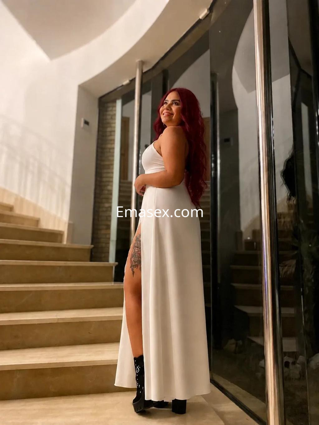 Escort en Valencia luce un vestido blanco largo, de pie en una escalera, con cabello largo y rojo, mostrando un tatuaje en la pierna.