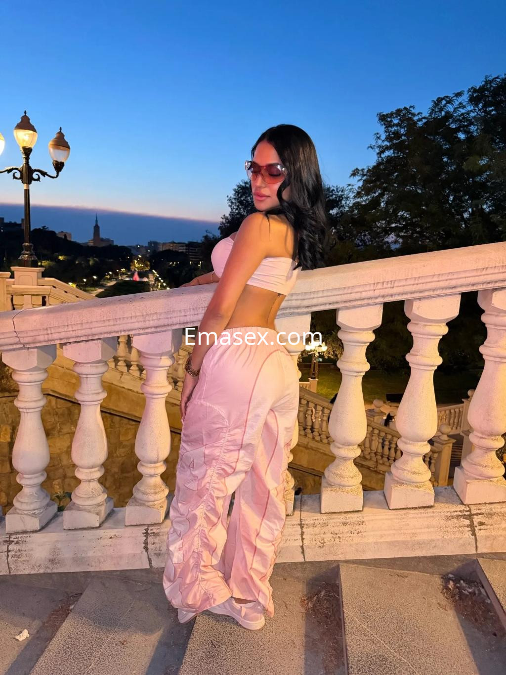 Puta en Zaragoza luce un conjunto rosa ajustado, posando con gafas de sol ante un paisaje al atardecer.