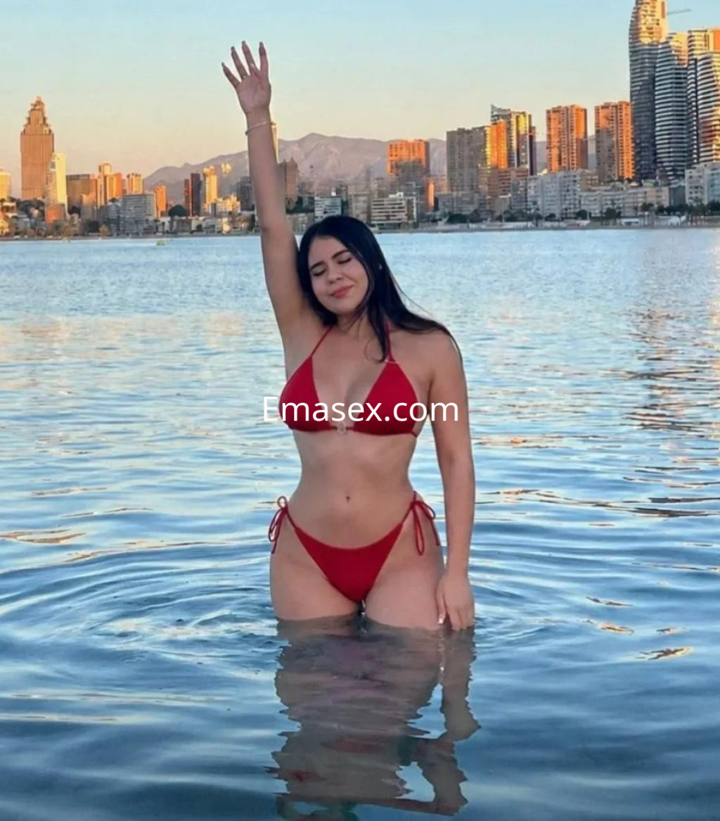 La puta está de pie en el agua con un bikini rojo, levantando un brazo, mientras se ve el skyline de Madrid al fondo.