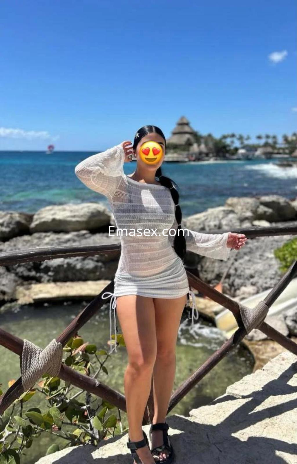 Escort en Alicante con vestido de malla blanca, posando frente al mar y un paisaje tropical.