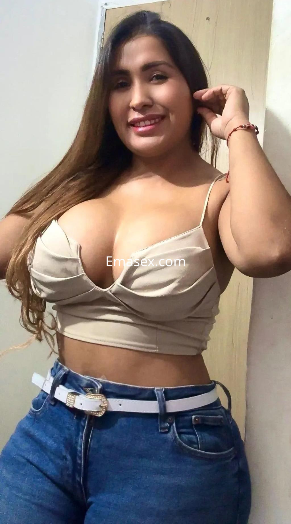 La escort en Alicante posando con una blusa beige ajustada y jeans de tiro alto, mostrando una sonrisa encantadora.