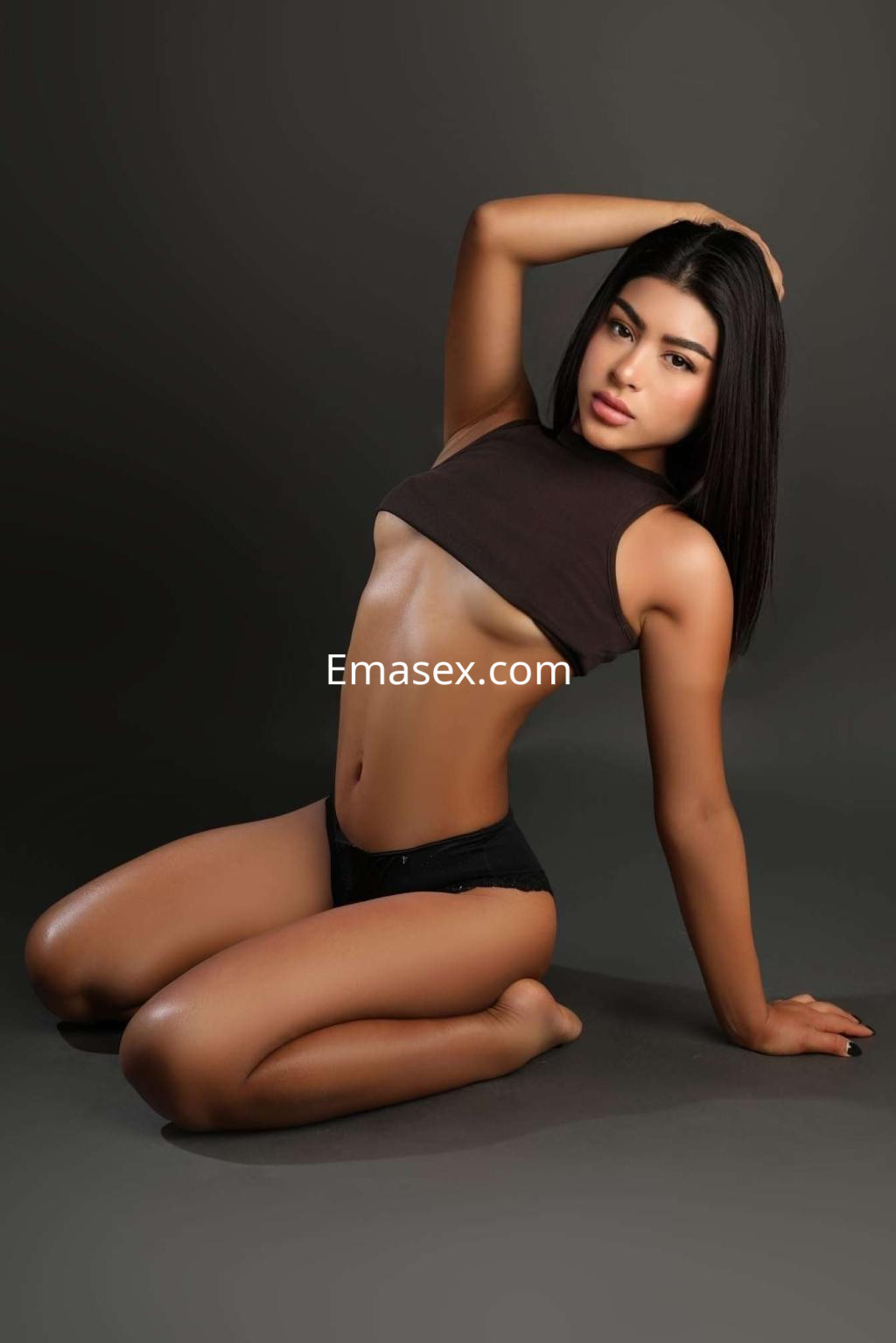 Puta de piel bronceada y cabello lacio, posando en Ibiza con top negro y tanga, sobre fondo gris.