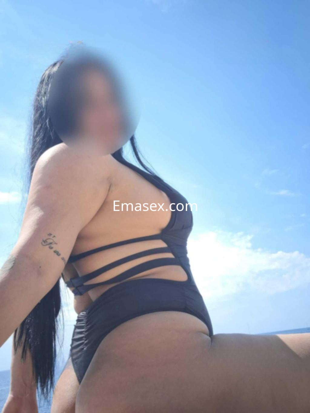 Escort en Barcelona con cabello largo y oscuro, posando en bikini negro bajo un cielo despejado.