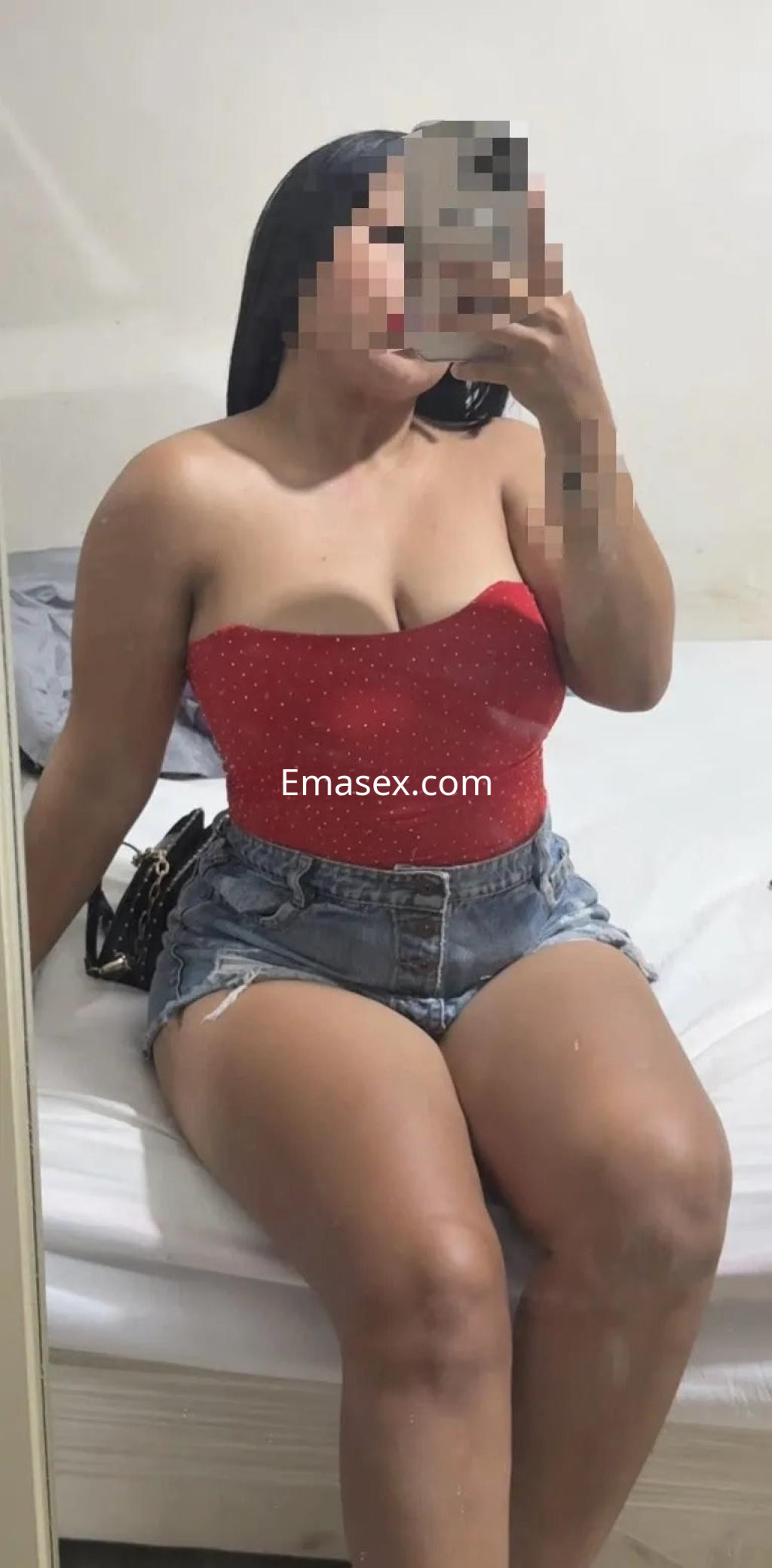 Escort en Córdoba sentada en una cama, viste un top rojo sin mangas y shorts de mezclilla, con cabello oscuro.