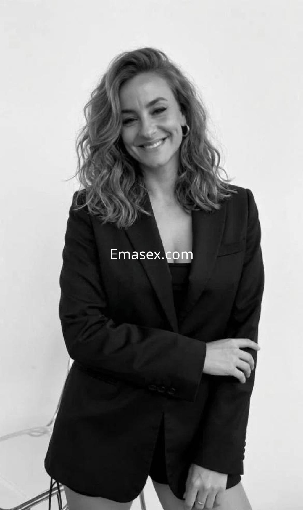 Escort en Lugo sonríe mientras lleva un blazer negro, con cabello rizado y un fondo claro.