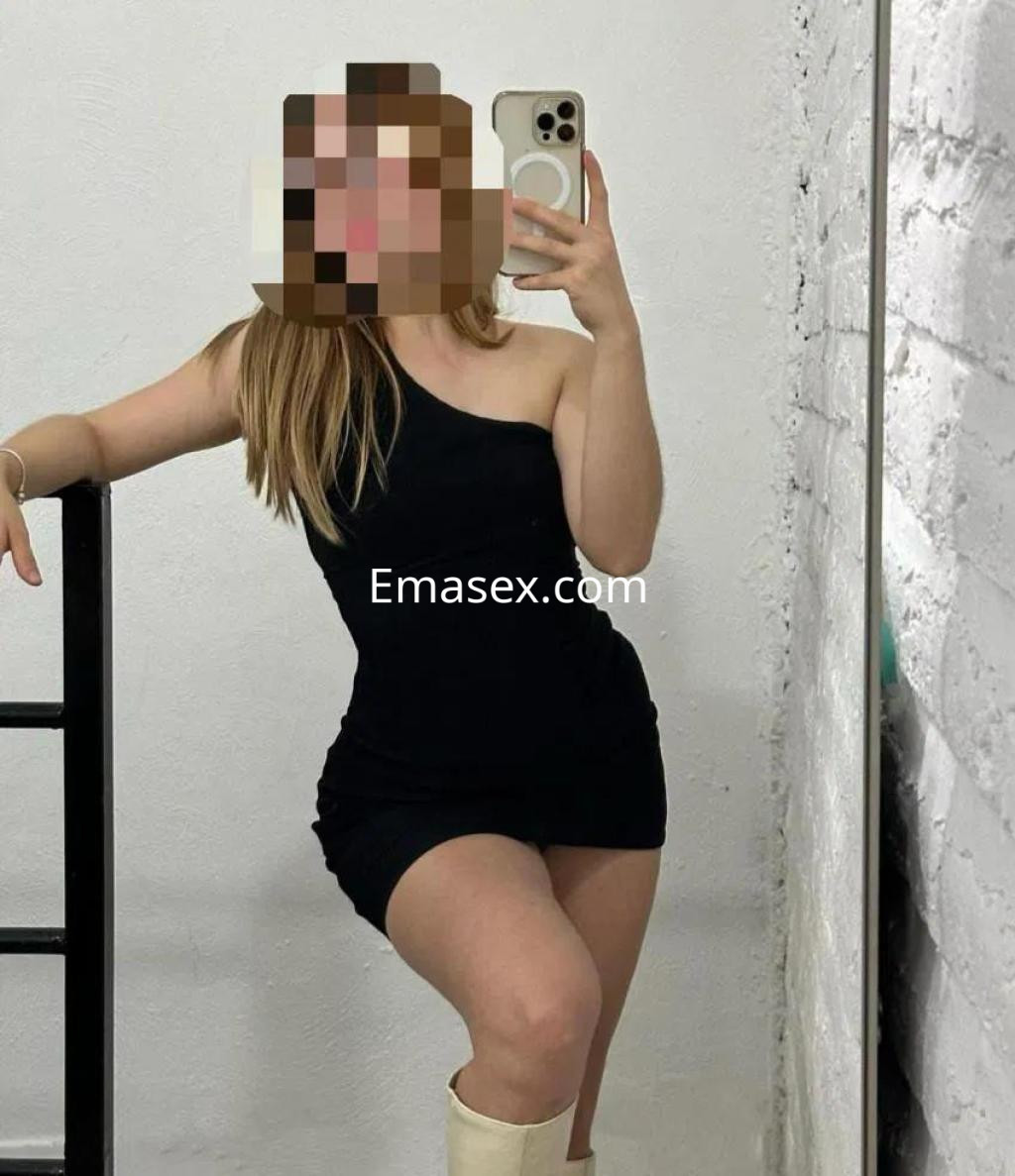 Escort en Las Palmas posando frente a un espejo, lleva un vestido negro de un solo hombro y botas blancas.