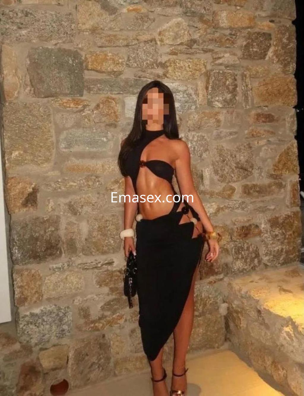 Escort en Las Palmas con un vestido negro que muestra su figura, posando frente a una pared de piedra.