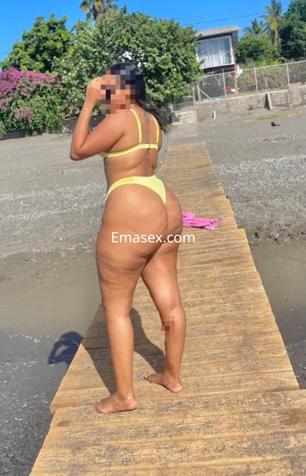 Escort de Pontevedra posando de espaldas en una pasarela de madera, luciendo un bikini amarillo en la playa.