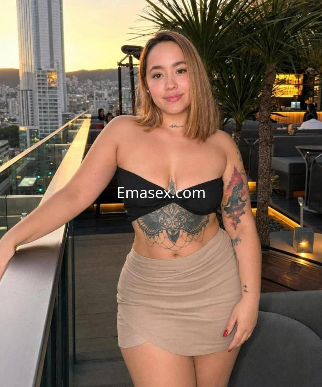 Escort en Toledo con cabello castaño claro, viste un top negro y una falda corta beige, posando en una terraza moderna.