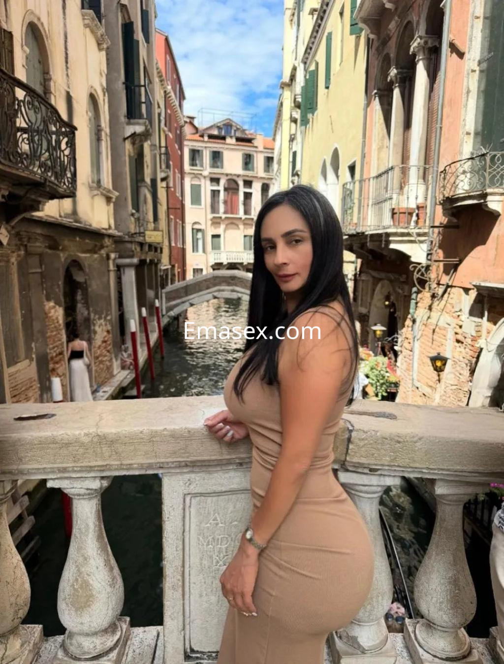 Escort en Valencia con vestido ajustado color beige, posando junto a un canal en un entorno arquitectónico atractivo.
