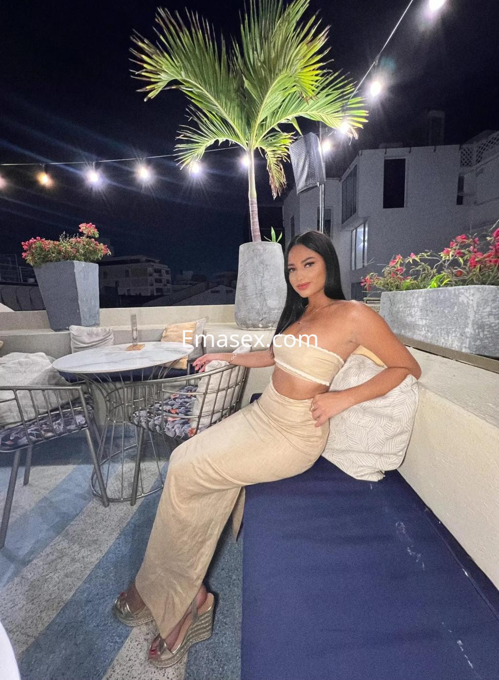 Puta sentada en un sofá en una terraza de Valencia, luciendo un top claro y una falda larga, con cabello lacio.