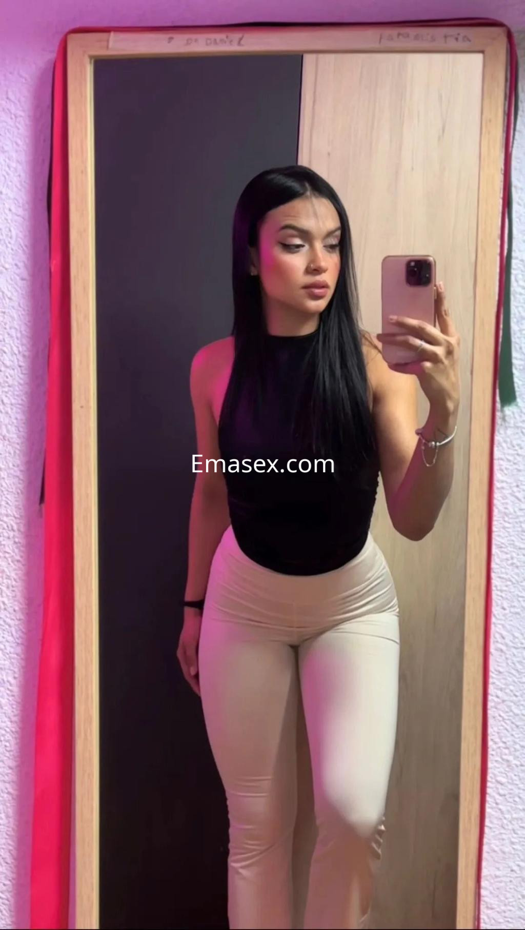Escort en Valencia con cabello lacio y oscuro, usando un top negro y pantalones ajustados claros frente a un espejo.