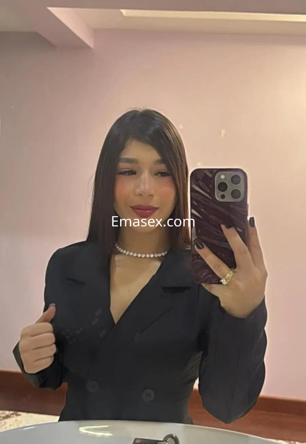Puta en Málaga viste un elegante traje negro con un collar de perlas, posando frente a un espejo.