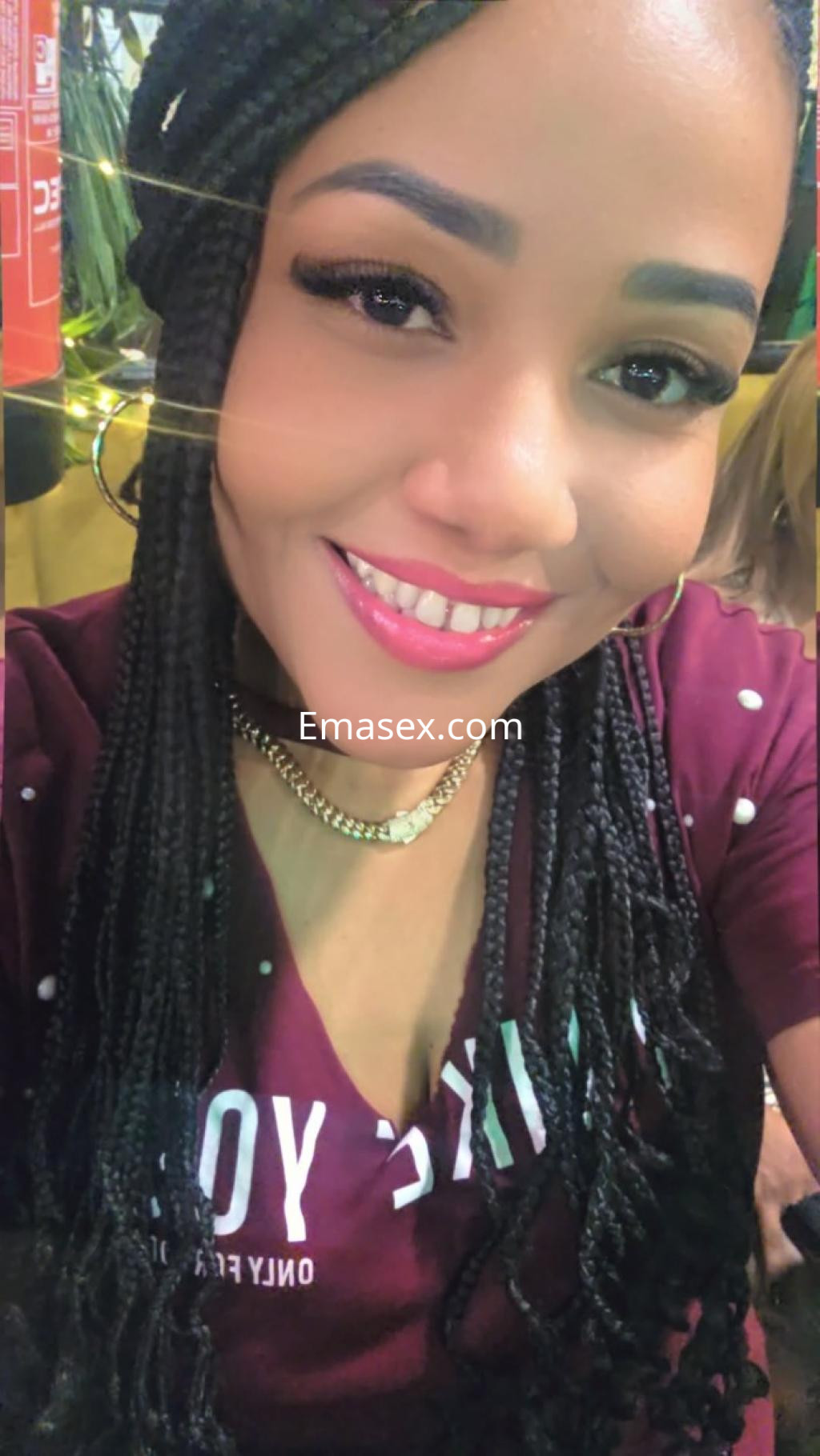 Escort en Gijón con cabello trenzado y blusa burdeos, sonríe mientras se toma un selfie en un ambiente colorido.