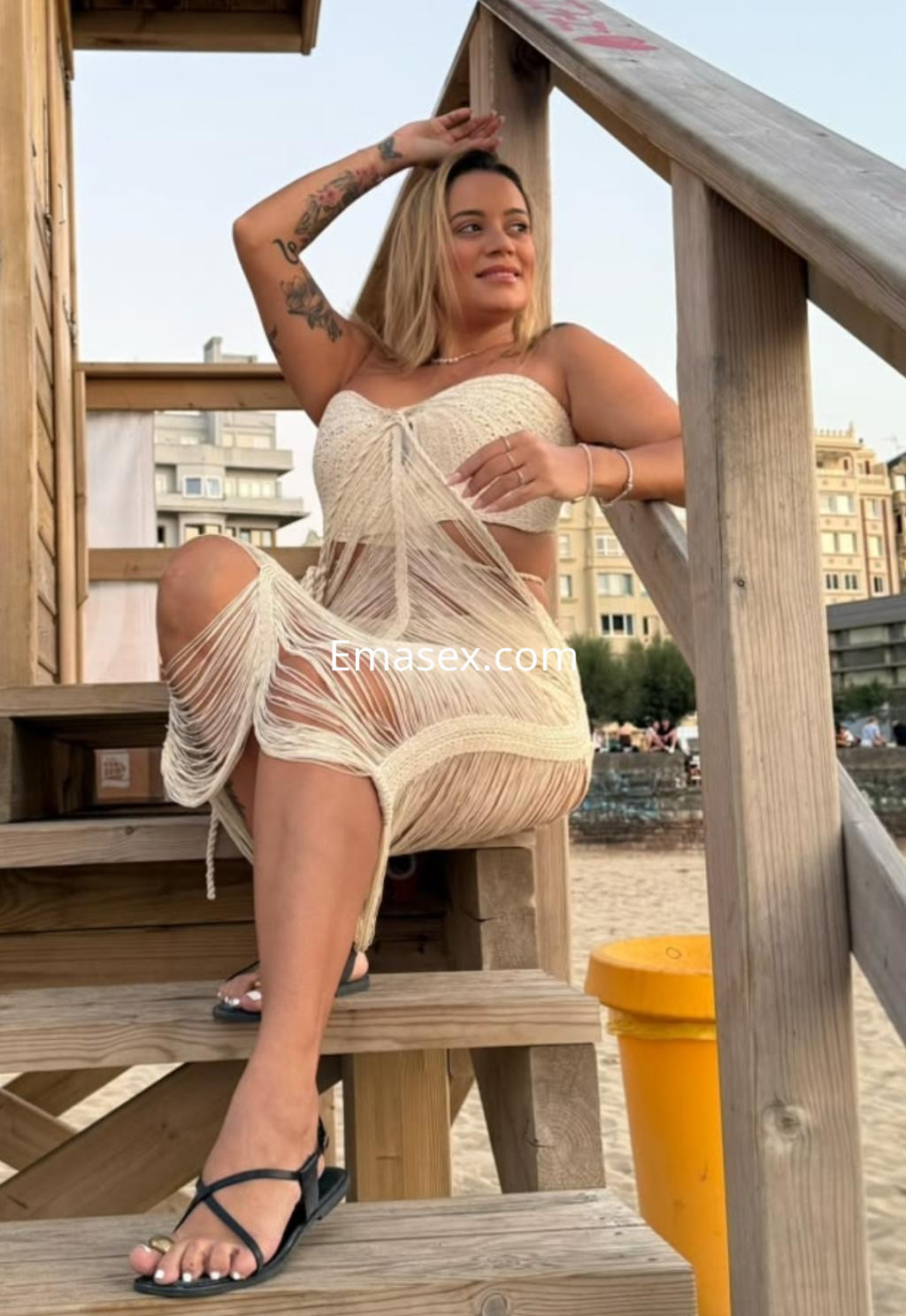 Escort en Donosti, sentada en una escalera de madera, vistiendo un vestido blanco semitransparente y sandalias.