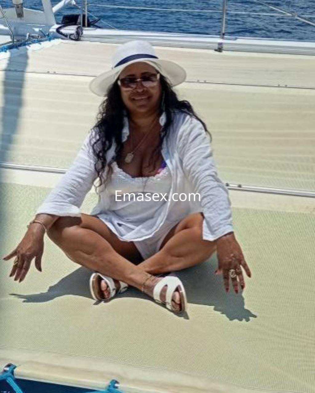 Escort en Las Palmas, sentada en un yate, con sombrero blanco, blusa ligera y sandalias, disfrutando del sol.