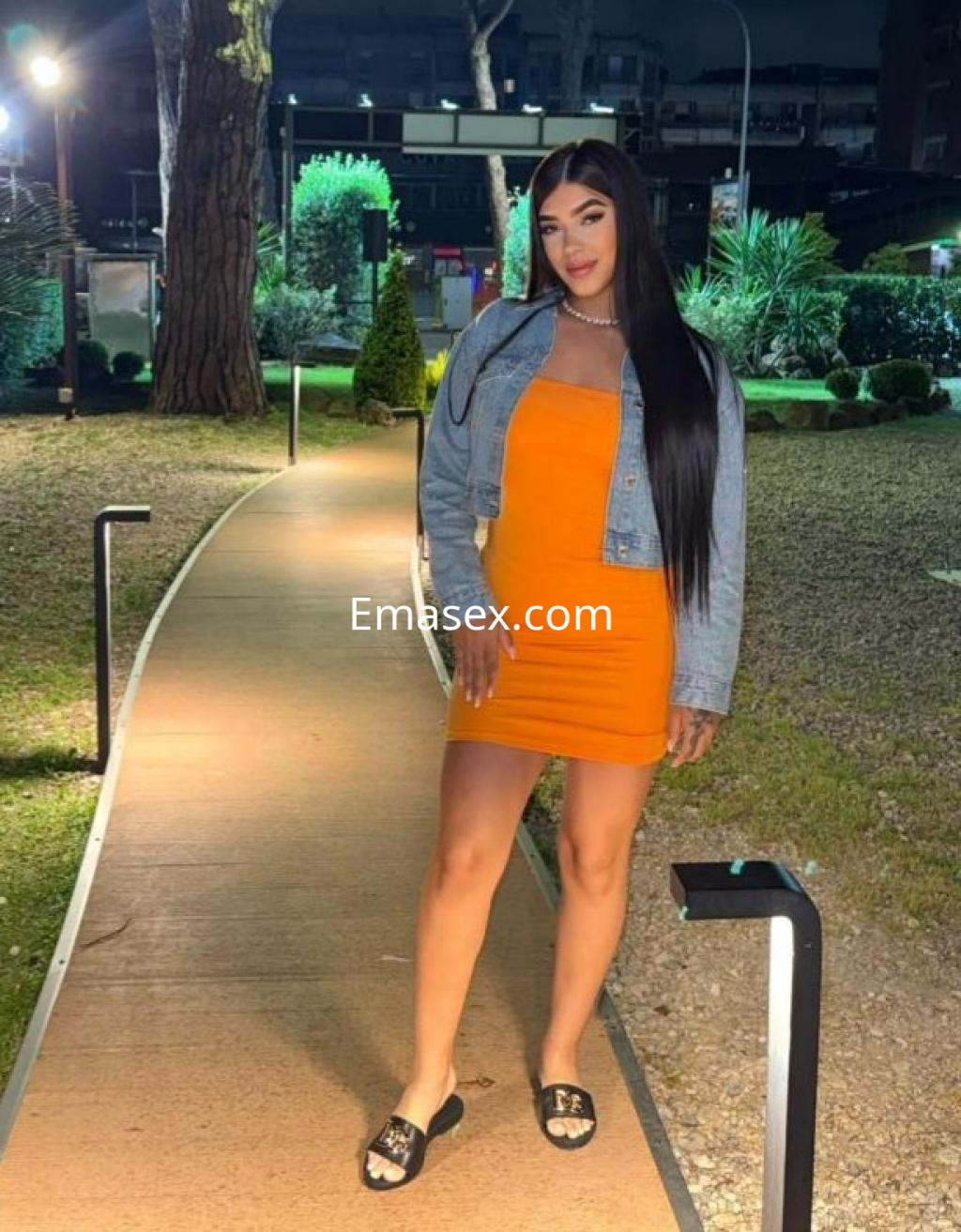 Escort en Las Palmas luciendo un vestido naranja ajustado y chaqueta de mezclilla, posando en un camino iluminado.
