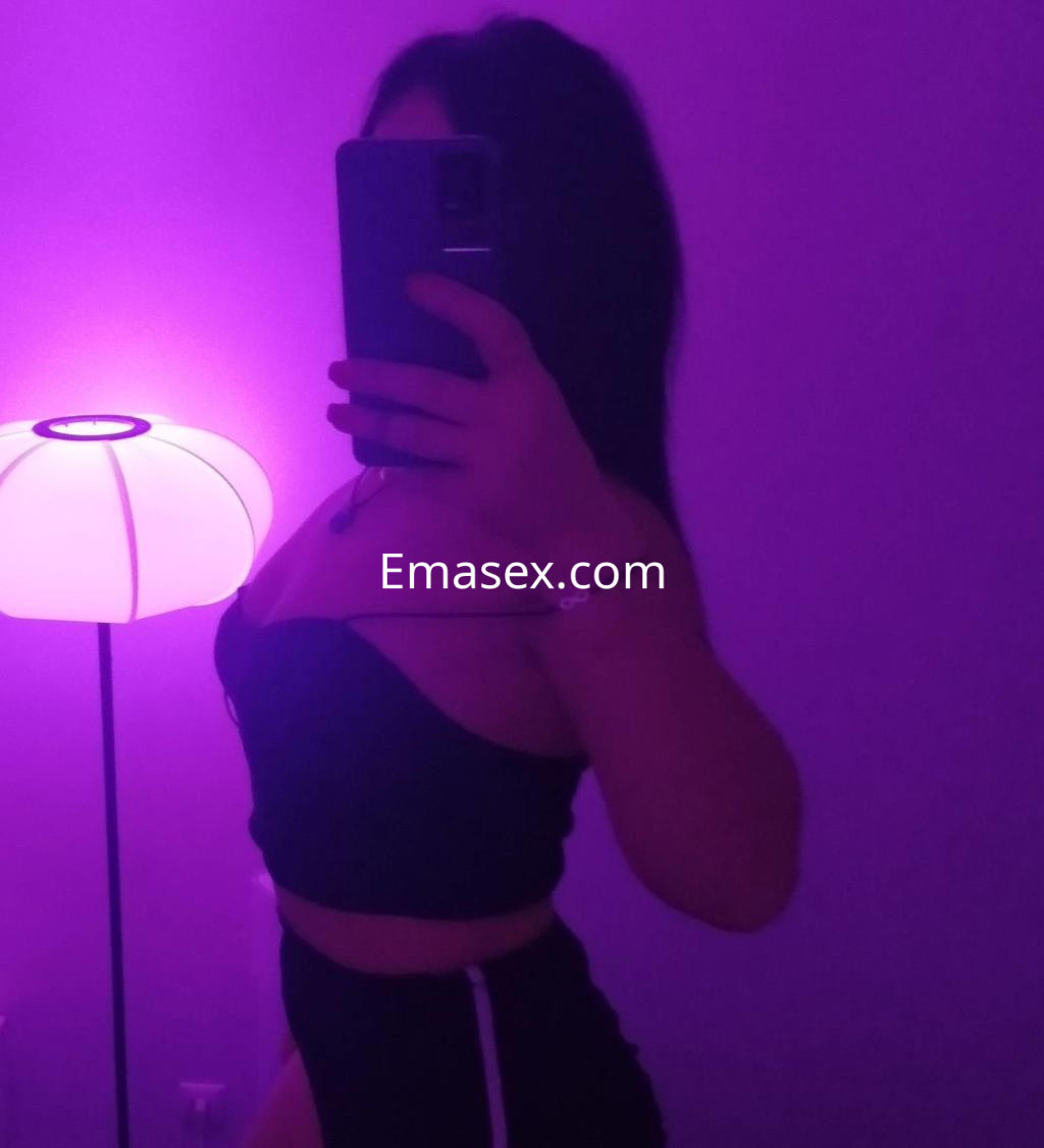 Escort en Córdoba posando de perfil con top negro y pantalones ajustados, en un ambiente iluminado con luz morada.