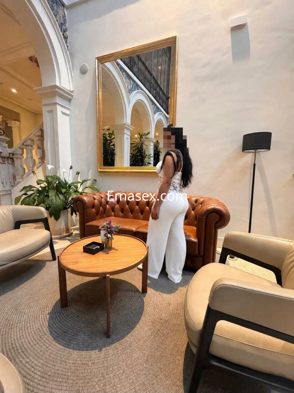 Escort en Granada, de pie en un elegante salón con sofá marrón, vistiendo un conjunto blanco, cabello largo y oscuro.