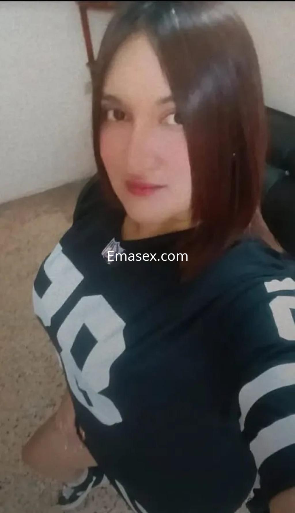 Escort en Granada con cabello castaño, viste una camiseta deportiva negra y shorts, sonriendo en un ambiente interior.