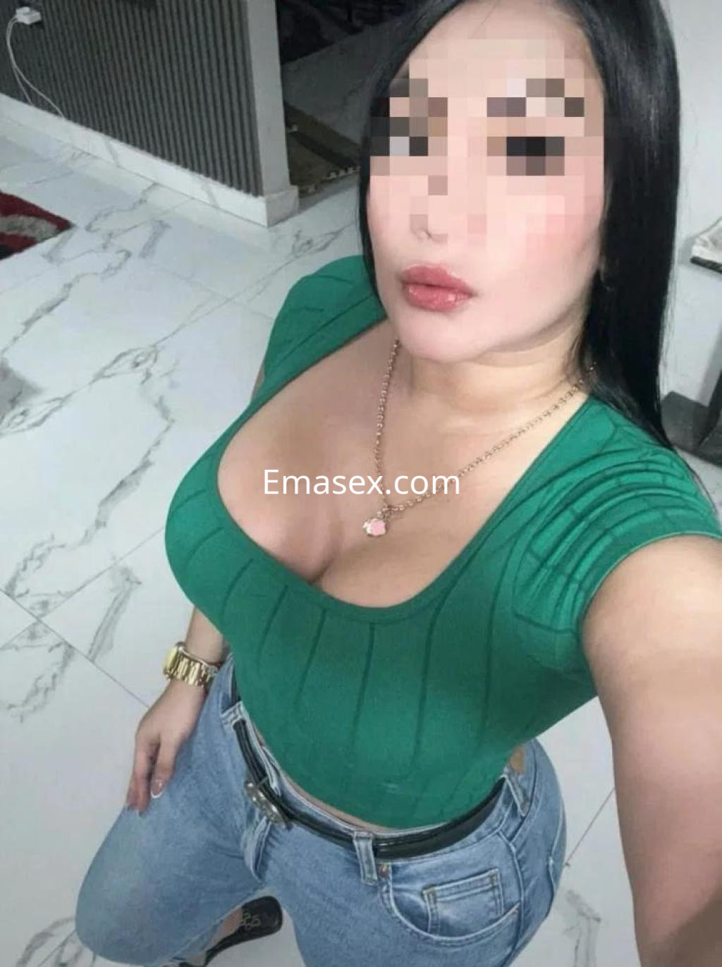 La puta muestra una pose seductora con una blusa verde ajustada y jeans, en un entorno interior elegante de León.