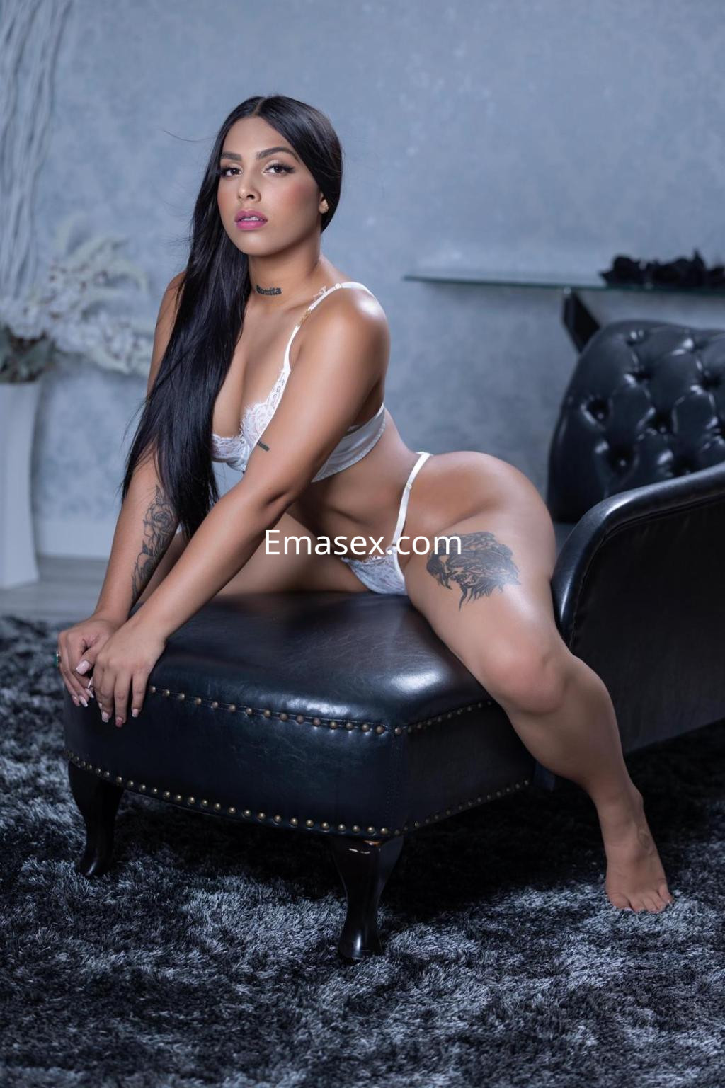 Escort en Madrid posando sobre un sofá negro, con lencería blanca, cabello largo y lacio, y tatuajes visibles.