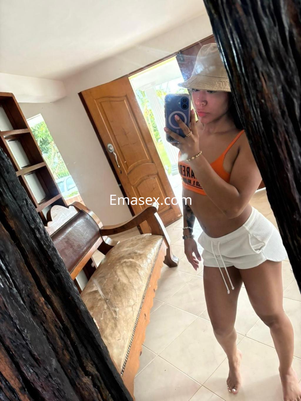 Escort en Santander se refleja en un espejo, vistiendo top naranja y pantalones cortos blancos, con un sombrero.