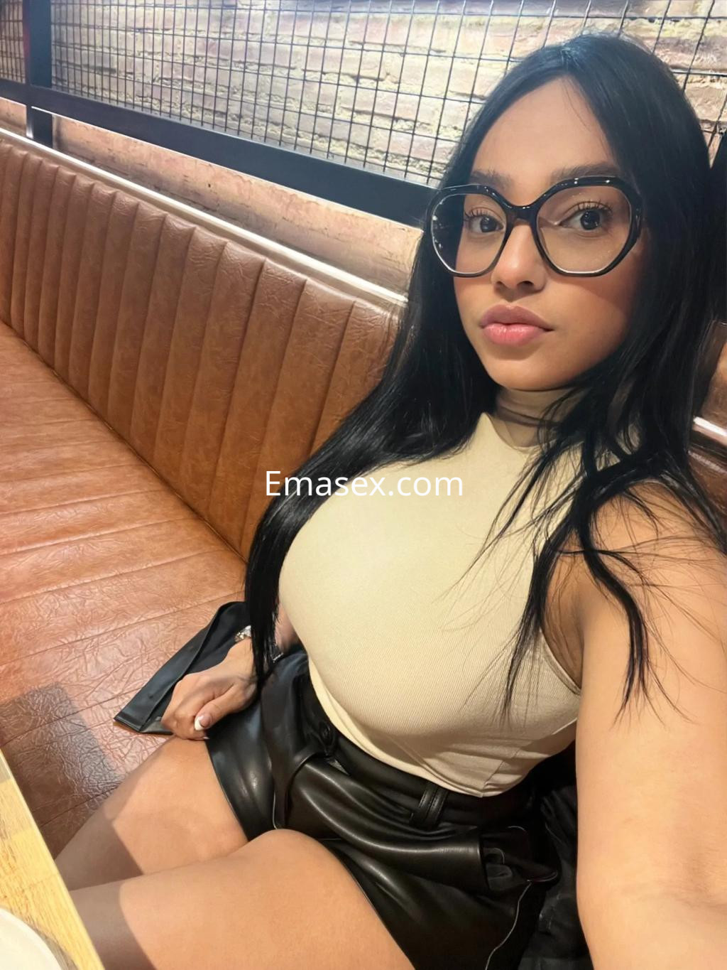 Escort en Vitoria, con cabello largo y oscuro, viste una blusa ajustada y una falda corta, posando en un restaurante.
