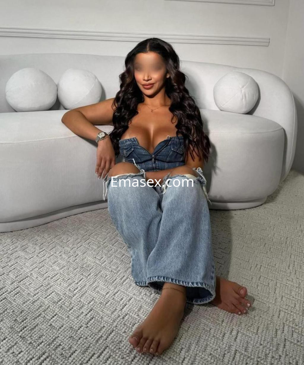 Escort en Albacete sentada en un sofá, con un top de mezclilla, jeans claros y cabello largo y ondulado.