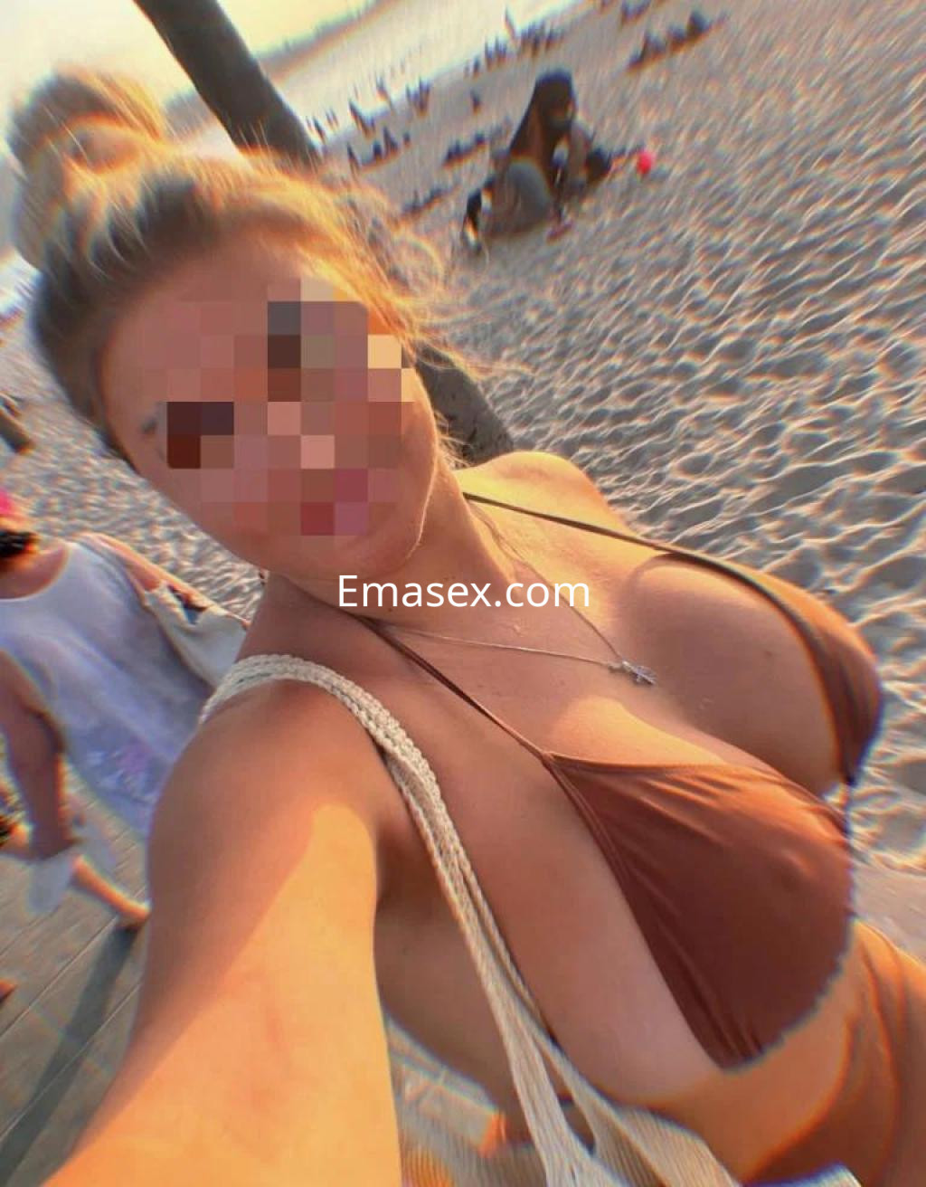 Escort en Almería, posando en la playa con un bikini marrón y una bolsa de playa, bajo la luz del atardecer.