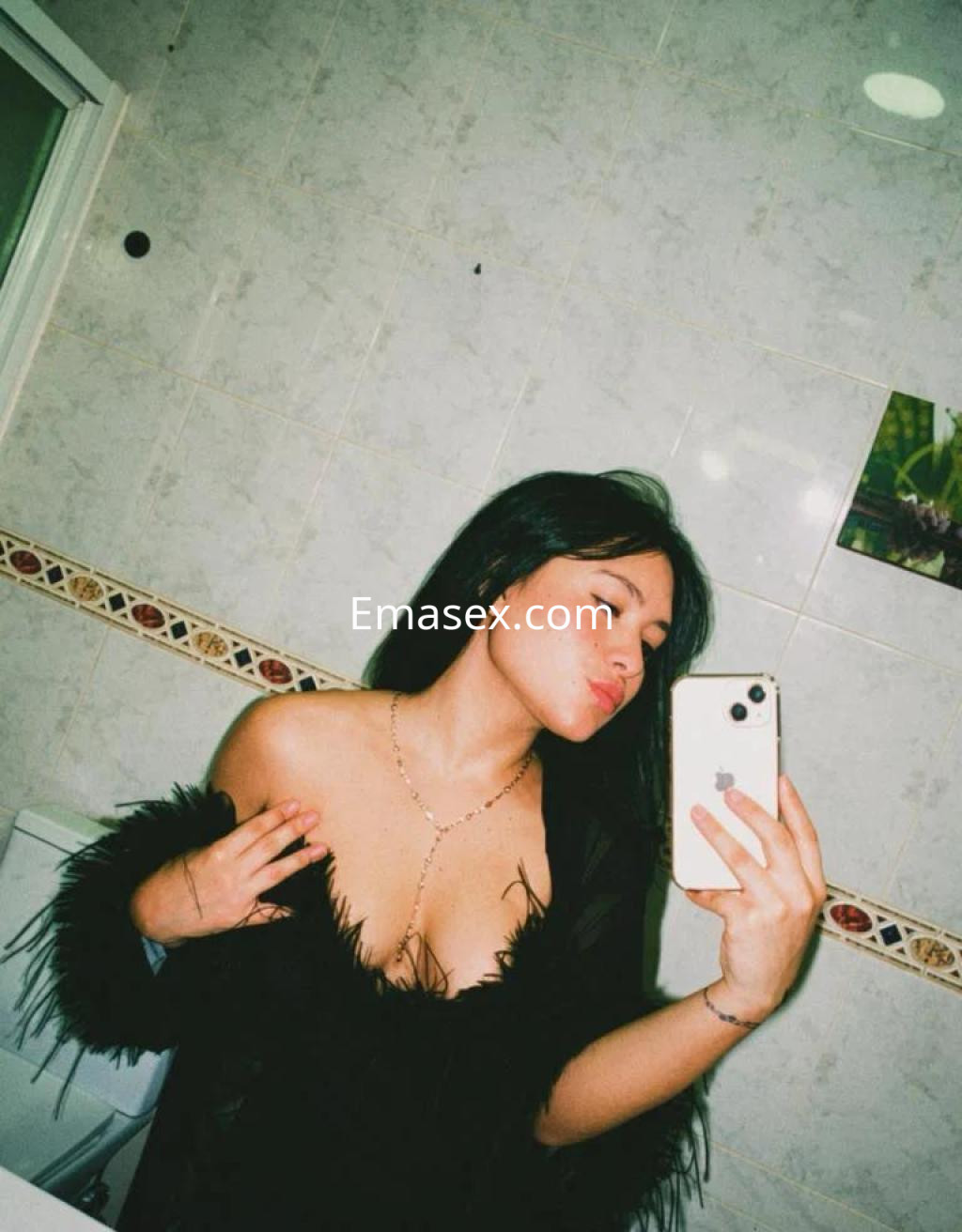 Escort en Sevilla se toma una selfie en un baño, vistiendo un top negro con detalles de plumas y luciendo un collar.