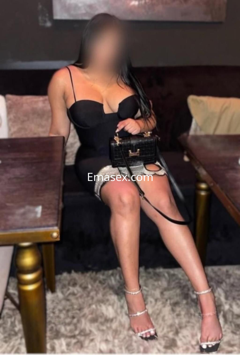 Escort en Bilbao luciendo un vestido negro ajustado, con tirantes delgados y sandalias brillantes, sentada en un sofá.