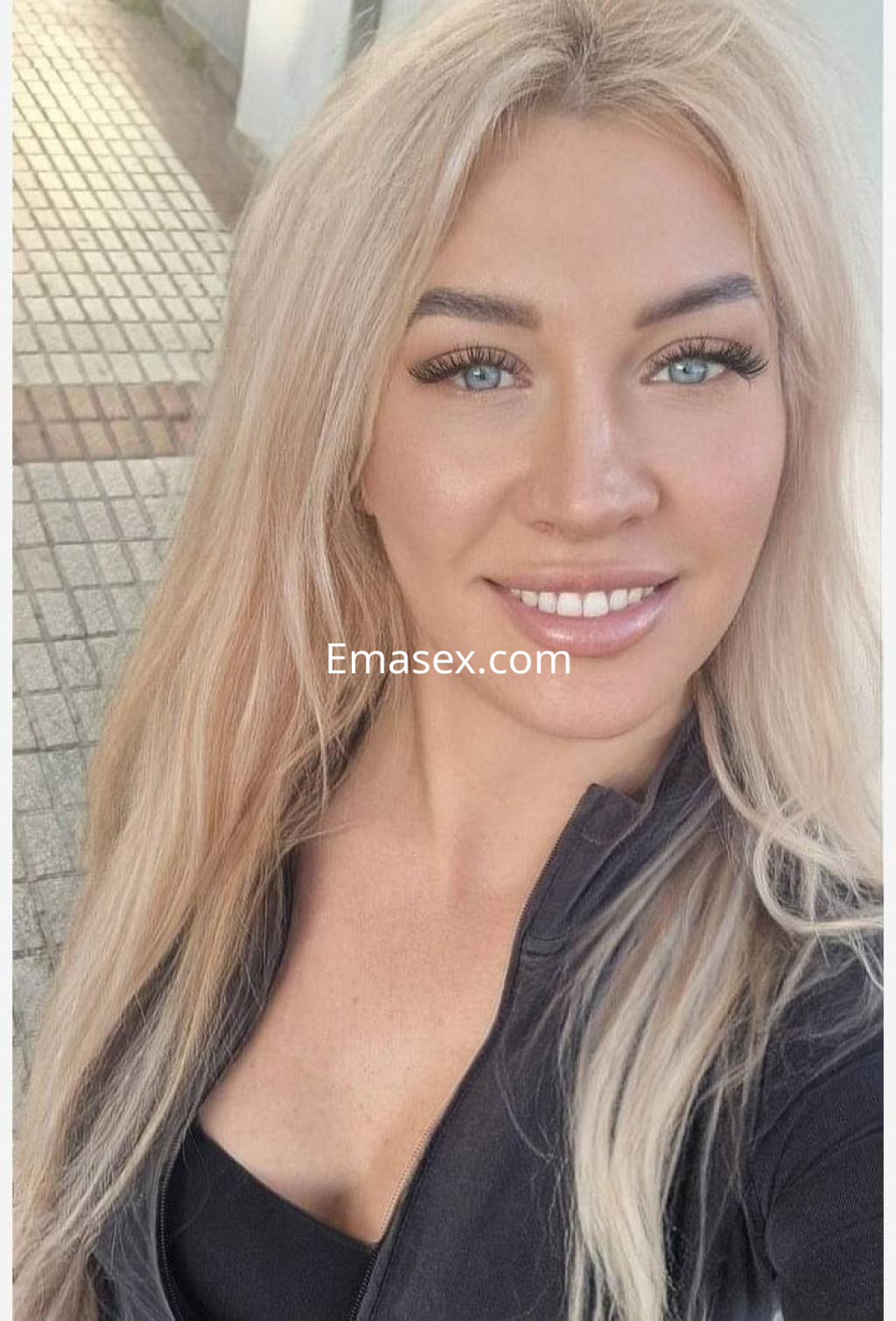 Escort en Bilbao, sonriendo a la cámara con cabello rubio largo y un top negro, en un entorno urbano iluminado.