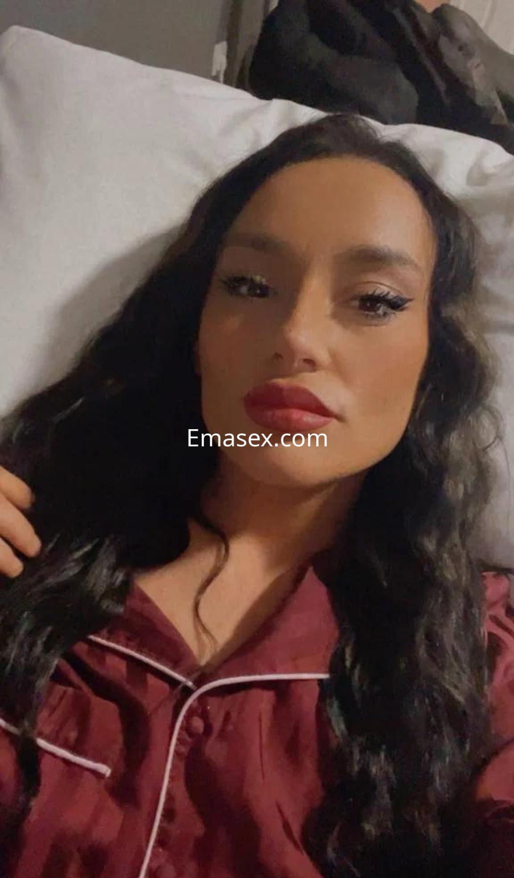 Escort en Girona con cabello largo y oscuro, luciendo pijama rojo, posando en una cama con una mirada seductora.