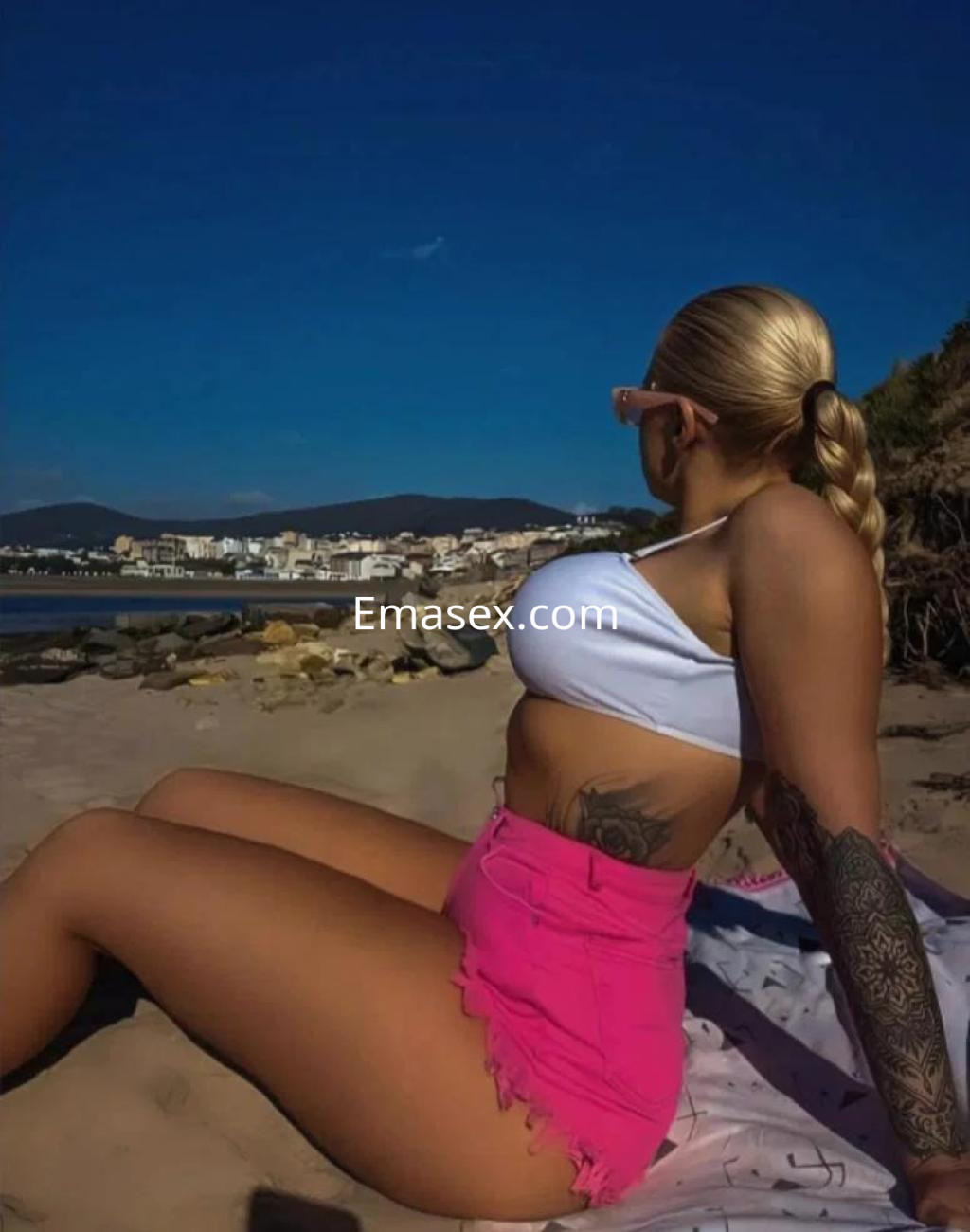 Escort en Lugo, con cabello rubio trenzado, vistiendo un top blanco y falda corta rosa, sentada en la playa.