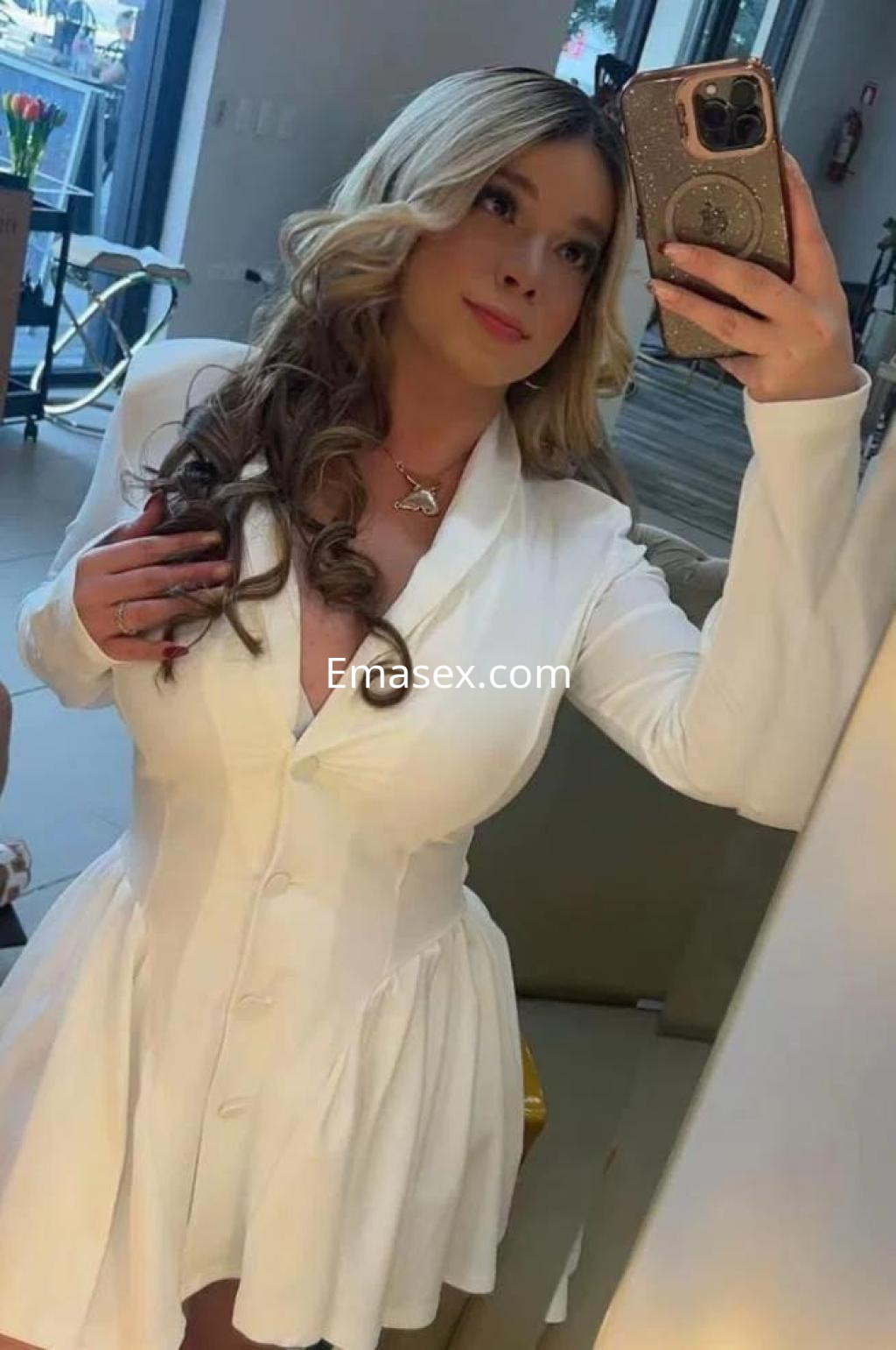 La escort en Málaga se retrata frente al espejo con un vestido blanco de corte ajustado y cabello rubio ondulado.