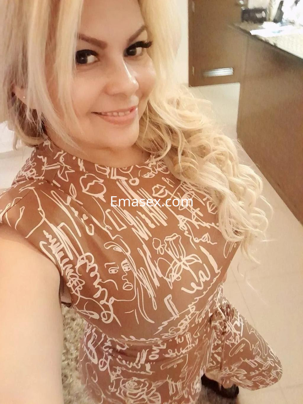 Puta sonriente con cabello rubio y rizado, vestida con un vestido marrón de estampado, en un entorno interior en Murcia.