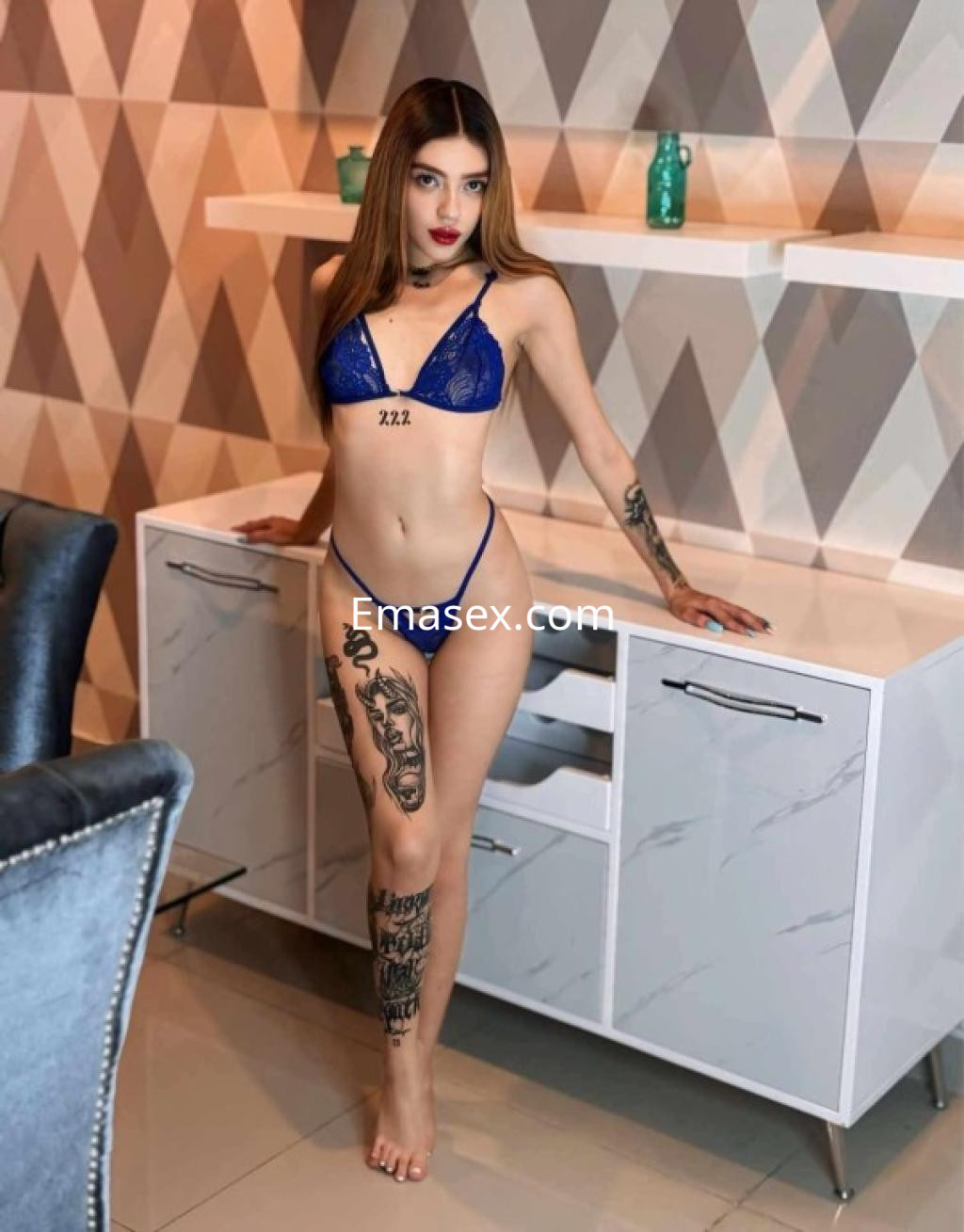 Escort en Tenerife, posando de pie junto a un mueble, viste un conjunto de lencería azul y exhibe tatuajes en su pierna.