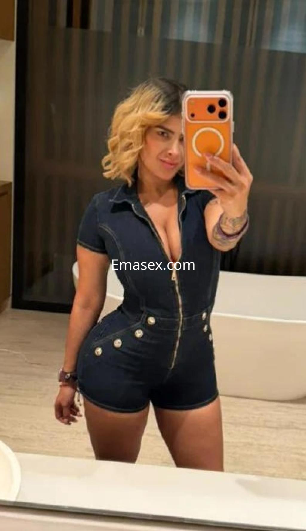 Escort en Málaga, con cabello rubio y rizado, viste un mono de mezclilla que realza su figura en un baño moderno.