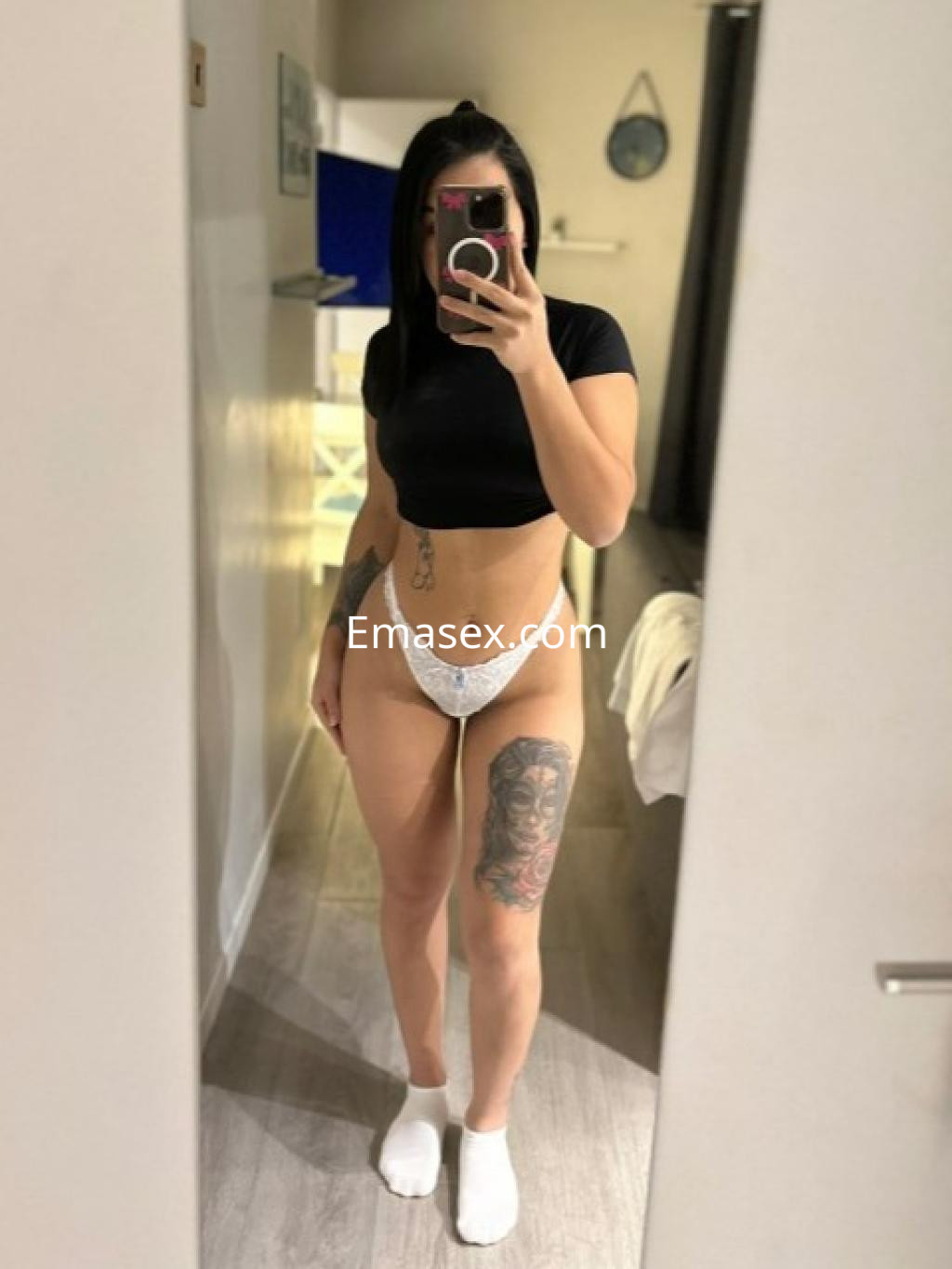 Escort en Castellón posando frente a un espejo, luciendo un top negro y una lencería blanca, con un tatuaje visible en la pierna.