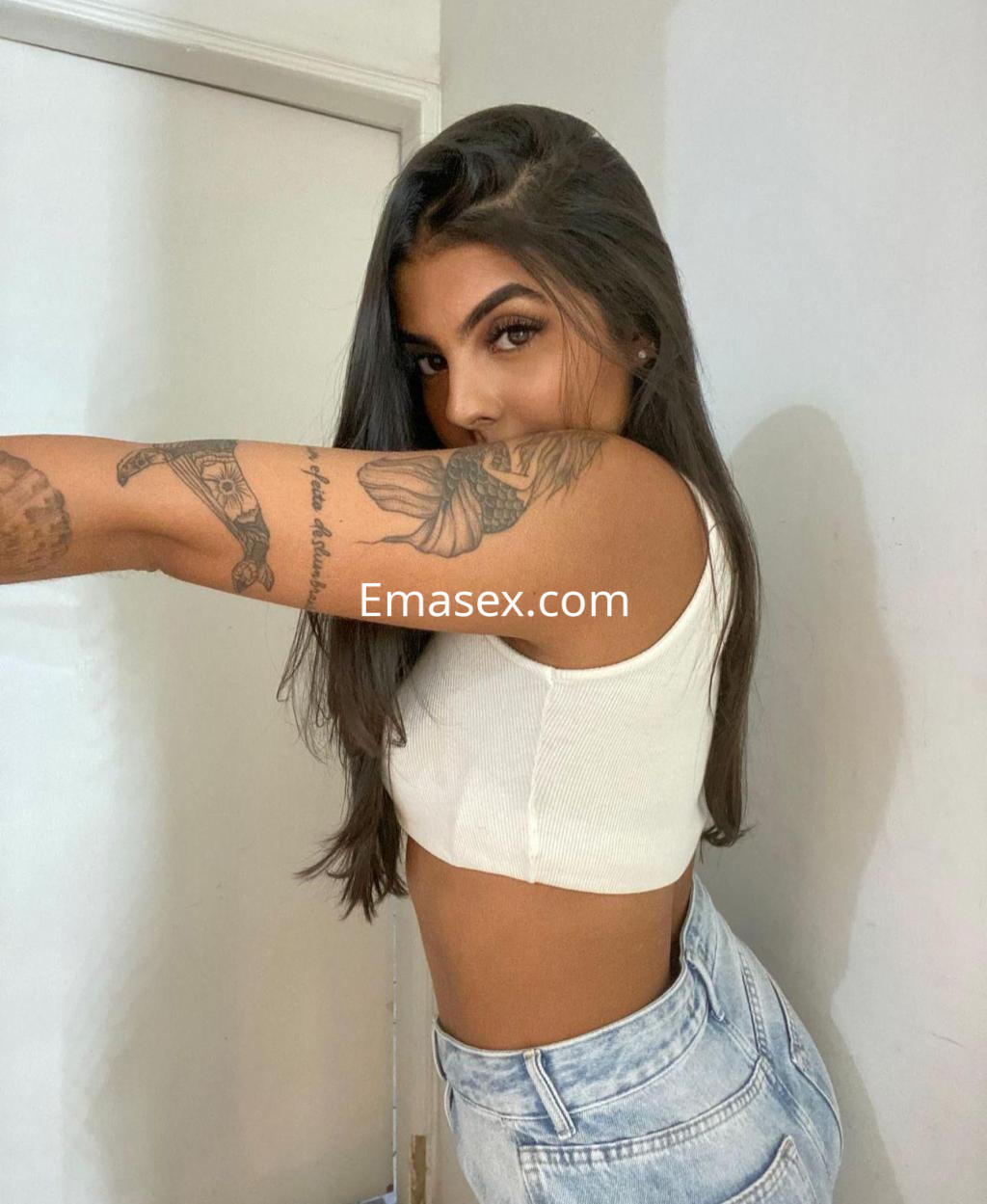 Escort en Sevilla con cabello largo y lacio, vistiendo un top blanco y shorts de mezclilla, posando de lado.
