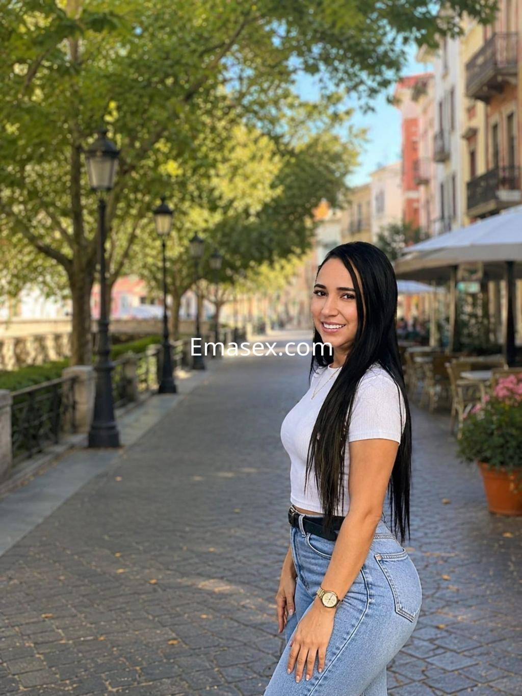 Escort en Bilbao con cabello largo y oscuro, viste una camiseta blanca y jeans ajustados, posando sonriendo en un paseo.