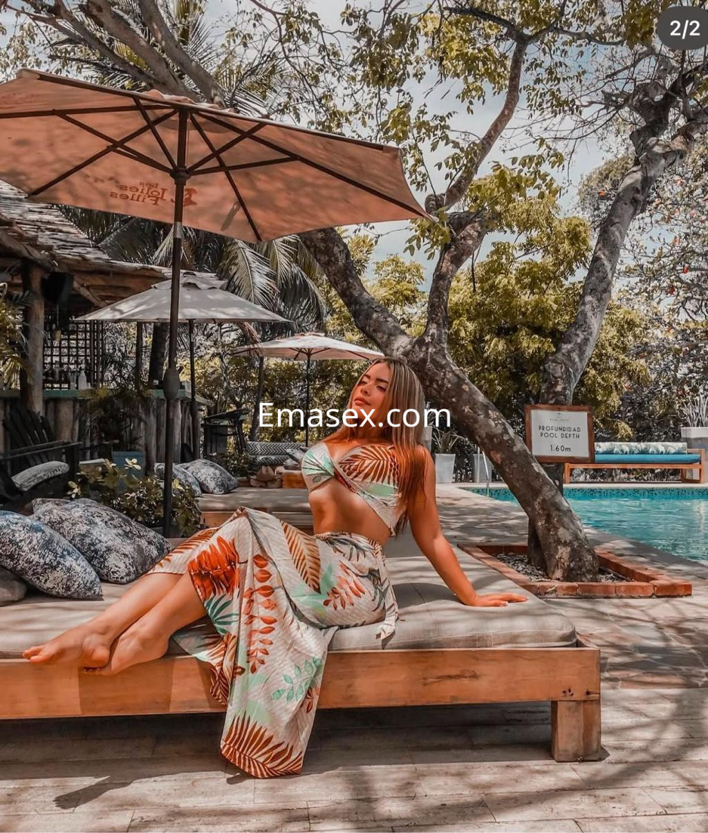 Escort en Irún sentada en una tumbona con un top y falda de estampado tropical, rodeada de vegetación y una piscina.