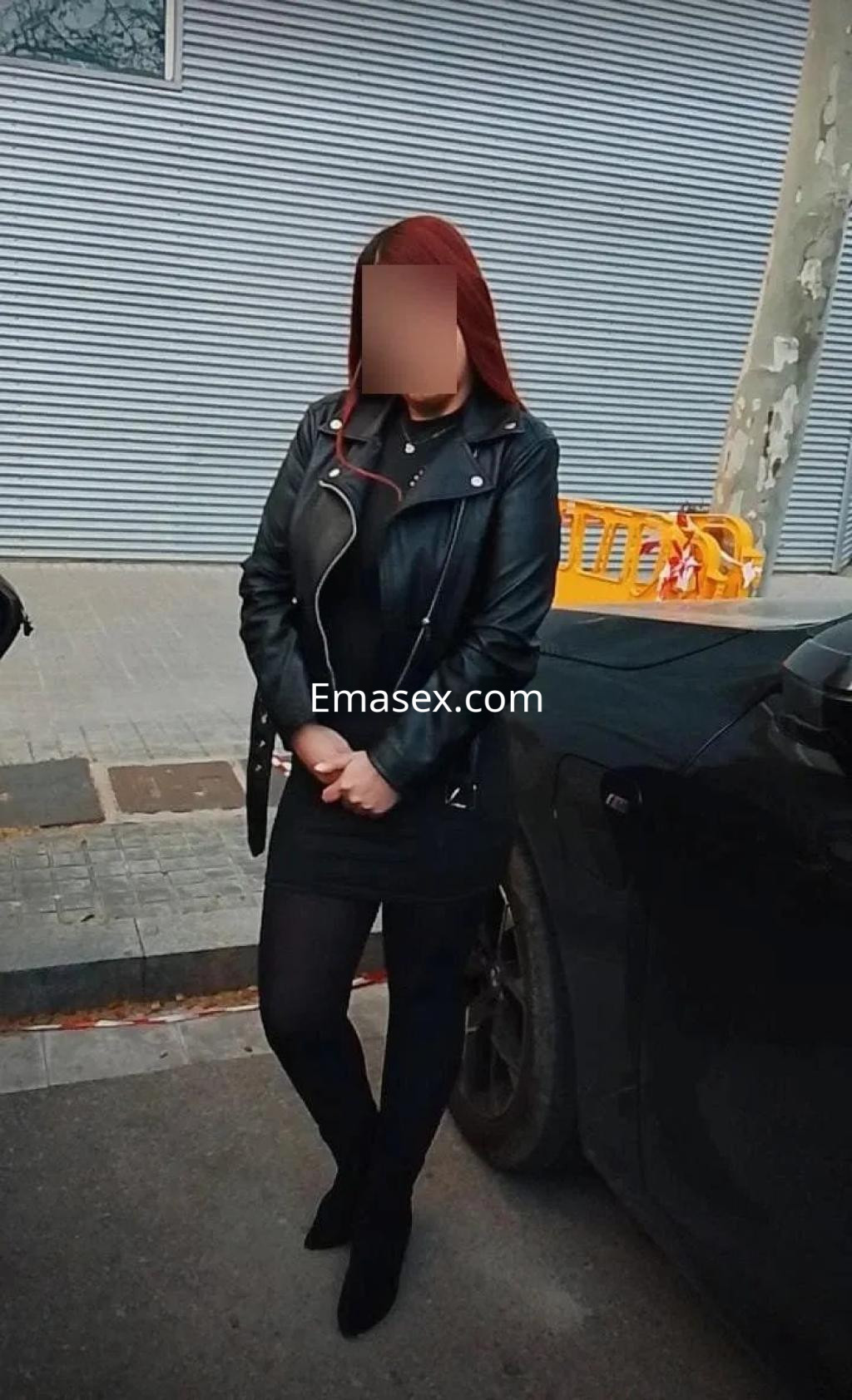 Puta de cabello castaño y rojizo, viste una chaqueta de cuero negra y se encuentra de pie junto a un coche en Barcelona.