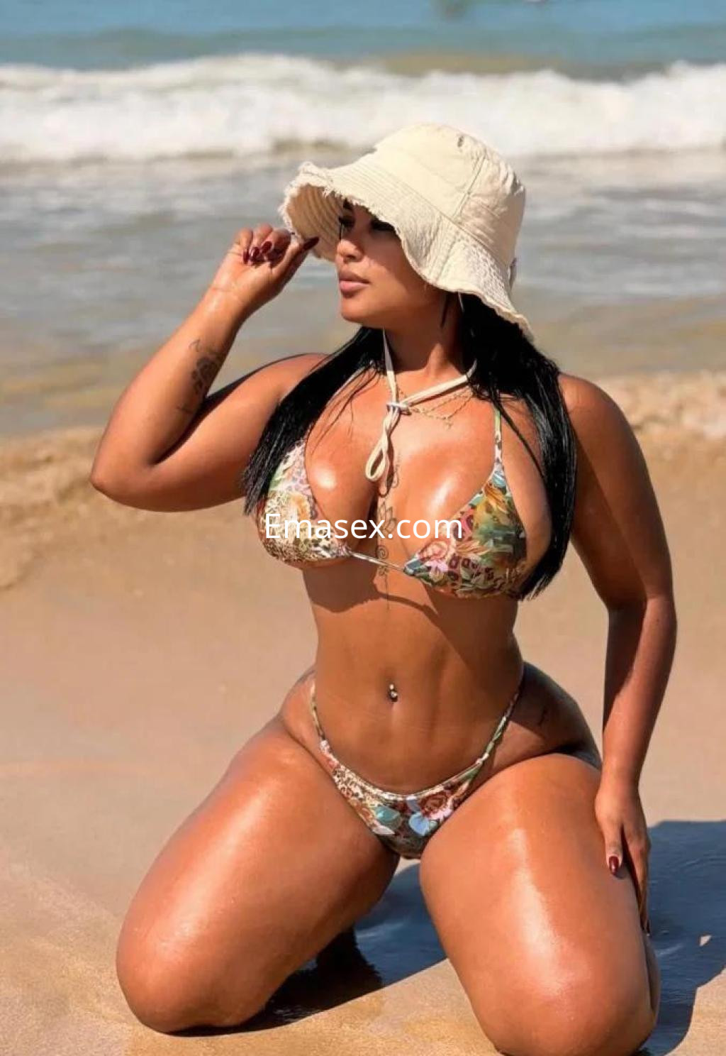 Escort de piel bronceada, posando en la playa de Barcelona con un bikini colorido y un sombrero blanco.