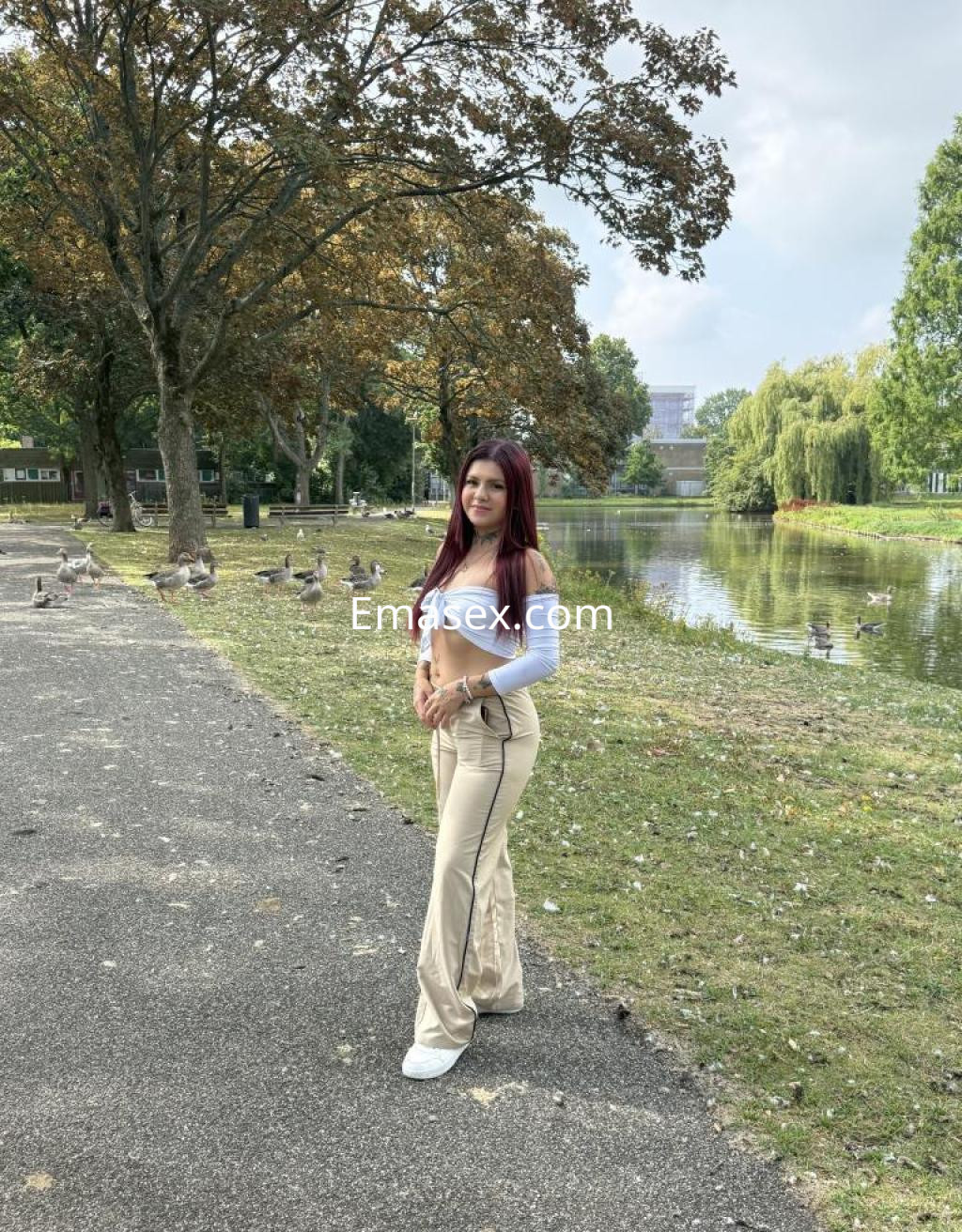 La puta está de pie junto a un lago en Cáceres, vistiendo una blusa blanca y pantalones claros, con cabello largo y castaño.
